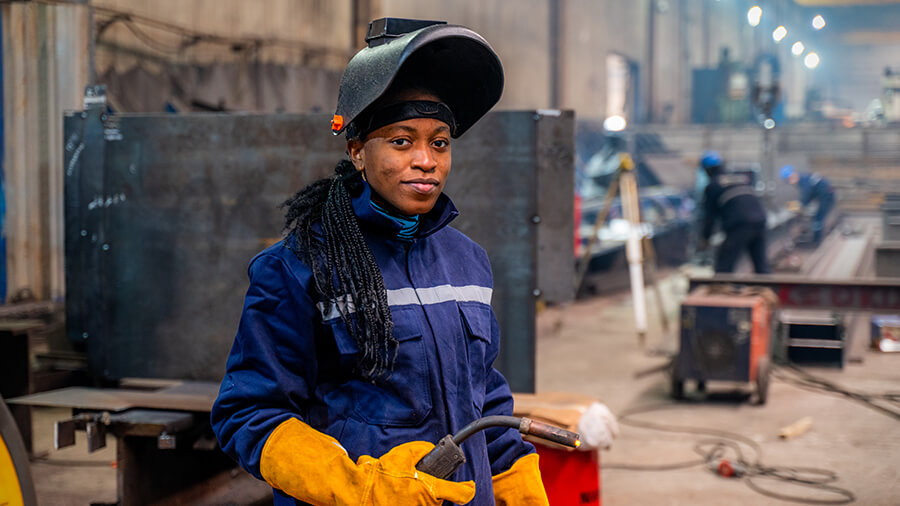 A welding student wears a helmet that's raised to show their face and gloves as they hold equipment.