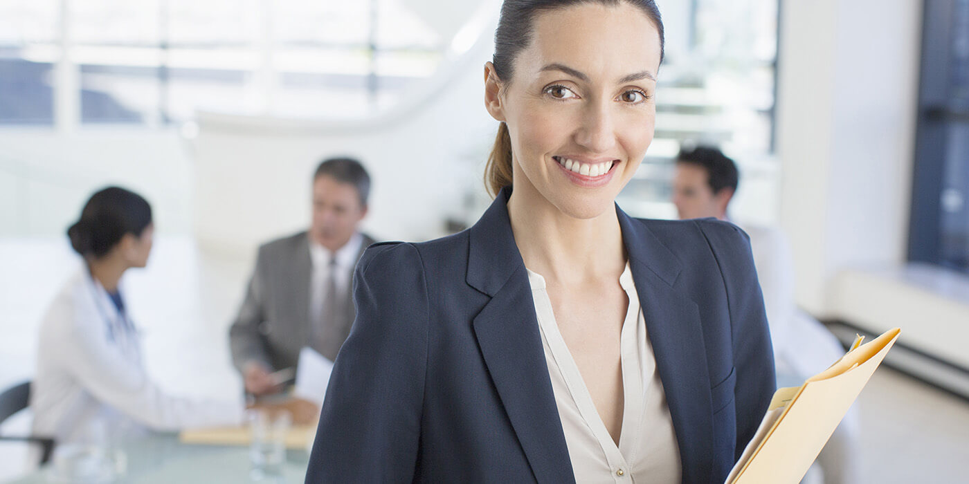 A woman in a blazer holds a couple of folders and smiles at the camera while colleagues meet in the background