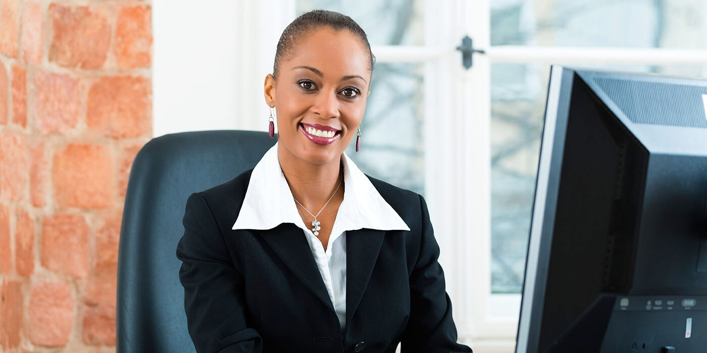 A woman in a business suit sits at a desk in front of a computer and smiles at the camera