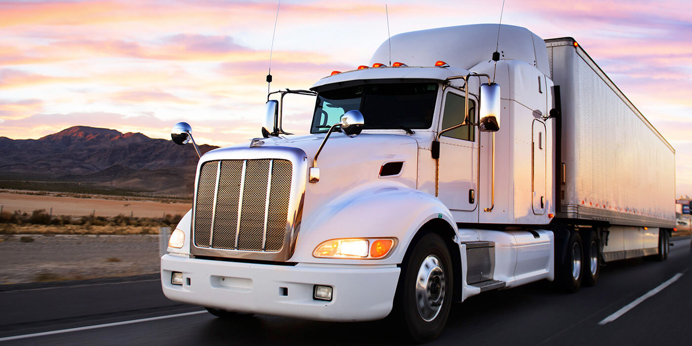 A close-up of a semi-truck driving on a highway with mountains and a sunset in the background