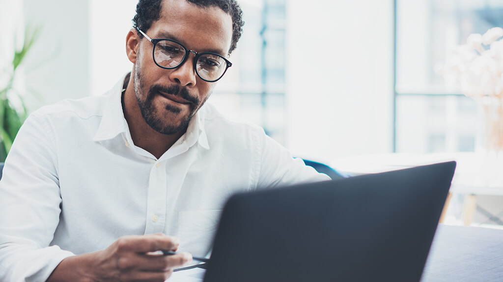 An adult in business casual clothing studies their laptop screen while holding a pen.