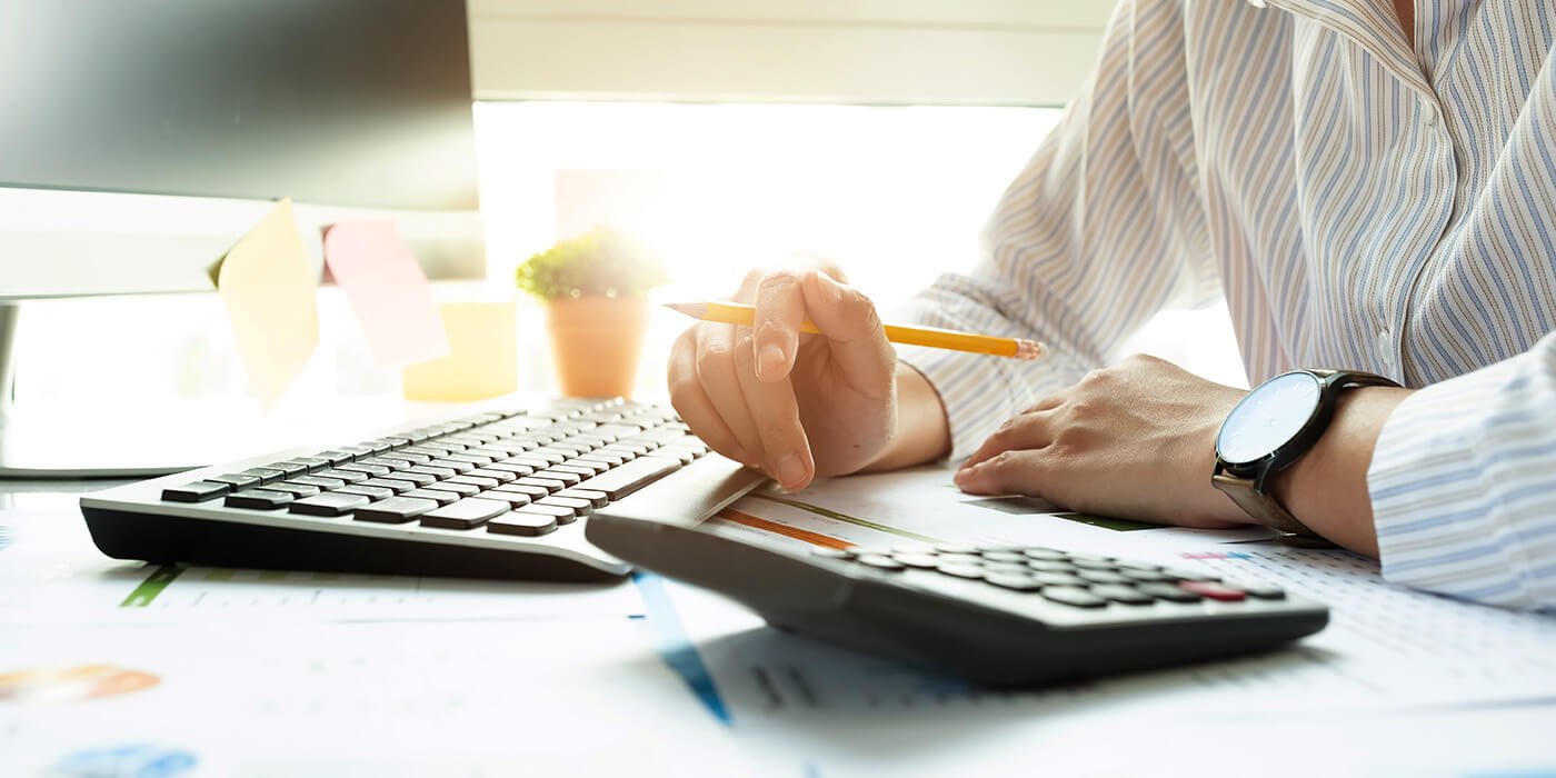 A close-up of hands above a calculator. A computer sits nearby. The person is wearing a white button-down shirt and a watch.